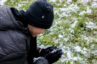 Genç çocuk kışın dışarıda ilk karla oynuyor. Sıcak ceket giyen ergen, şapkalı ve eldivenli genç yeşil çimlerde taze kara dokunuyor. Soğuk havada sezonluk gençlik anıları.
