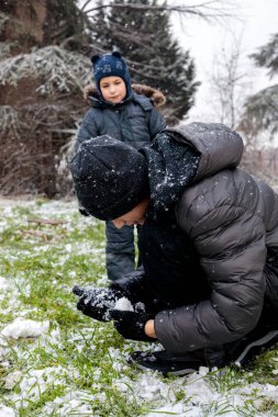 Genç çocuk çimlerin üzerinde çömelip kar şekillendirirken küçük çocuk kış karlarını seyrediyor. Kardeşler açık havada ilk karla oynuyor. Yerleşim bölgesinde doğal kış yaşam tarzı.