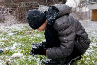 Kış karları çimenlerin üzerinde çömelmiş ve şekillendirilmiş genç çocuk, kalın ceketli ve bereli ergen çocuk açık havada ilk karla oynuyor, doğal kış yaşam tarzı.