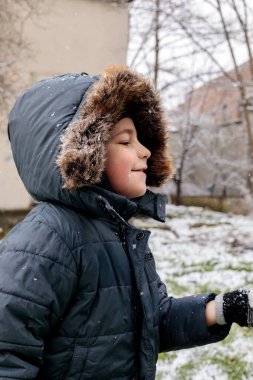 Kışın açık havada ilk kar yağışının keyfini çıkaran çocuk, kürk kapüşonlu ve eldivenli kalın bir ceket giyiyor, parkta gülümsüyor ve kar tanelerini yakalıyor, soğuk havada doğal çocukluk anı.