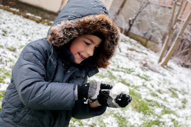Kışın açık havada ilk kar yağışının keyfini çıkaran çocuk, kürk kapüşonlu ve eldivenli kalın bir ceket giyiyor, parkta gülümsüyor ve kar tanelerini yakalıyor, soğuk havada doğal çocukluk anı.