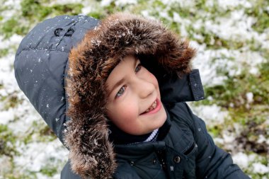 Kar yağışı sırasında dışarıda gülümseyen tüylü ceketli mutlu çocuk, soğuk havada ilk karın tadını çıkaran çocuğun portresi, mevsimsel kış çocukluğu konsepti.