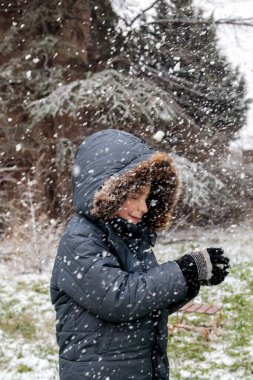 Kışın açık havada ilk kar yağışının keyfini çıkaran çocuk, kürk kapüşonlu ve eldivenli kalın bir ceket giyiyor, parkta gülümsüyor ve kar tanelerini yakalıyor, soğuk havada doğal çocukluk anı.