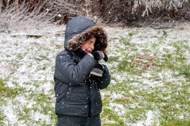 Kışın açık havada ilk kar yağışının keyfini çıkaran çocuk, kürk kapüşonlu ve eldivenli kalın bir ceket giyiyor, parkta gülümsüyor ve kar tanelerini yakalıyor, soğuk havada doğal çocukluk anı.
