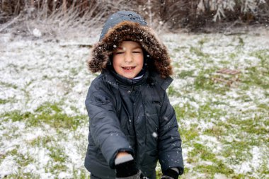 Kışın açık havada ilk kar yağışının keyfini çıkaran çocuk, kürk kapüşonlu ve eldivenli kalın bir ceket giyiyor, parkta gülümsüyor ve kar tanelerini yakalıyor, soğuk havada doğal çocukluk anı.