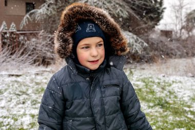 Kışın açık havada ilk kar yağışının keyfini çıkaran çocuk, kürk kapüşonlu ve eldivenli kalın bir ceket giyiyor, parkta gülümsüyor ve kar tanelerini yakalıyor, soğuk havada doğal çocukluk anı.