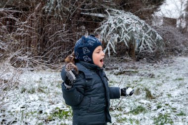 Kışın açık havada ilk kar yağışının keyfini çıkaran çocuk, kürk kapüşonlu ve eldivenli kalın bir ceket giyiyor, parkta gülümsüyor ve kar tanelerini yakalıyor, soğuk havada doğal çocukluk anı.
