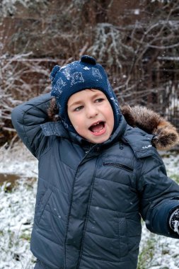 Kışın açık havada ilk kar yağışının keyfini çıkaran çocuk, kürk kapüşonlu ve eldivenli kalın bir ceket giyiyor, parkta gülümsüyor ve kar tanelerini yakalıyor, soğuk havada doğal çocukluk anı.