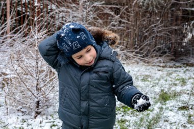 Kışın açık havada ilk kar yağışının keyfini çıkaran çocuk, kürk kapüşonlu ve eldivenli kalın bir ceket giyiyor, parkta gülümsüyor ve kar tanelerini yakalıyor, soğuk havada doğal çocukluk anı.