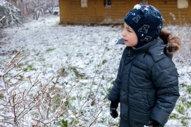 Kışın açık havada ilk kar yağışının keyfini çıkaran çocuk, kürk kapüşonlu ve eldivenli kalın bir ceket giyiyor, parkta gülümsüyor ve kar tanelerini yakalıyor, soğuk havada doğal çocukluk anı.