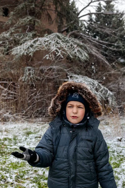 Kışın açık havada ilk kar yağışının keyfini çıkaran çocuk, kürk kapüşonlu ve eldivenli kalın bir ceket giyiyor, parkta gülümsüyor ve kar tanelerini yakalıyor, soğuk havada doğal çocukluk anı.