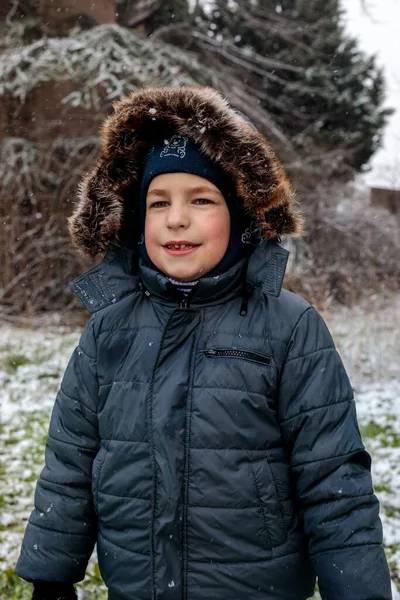 Kışın açık havada ilk kar yağışının keyfini çıkaran çocuk, kürk kapüşonlu ve eldivenli kalın bir ceket giyiyor, parkta gülümsüyor ve kar tanelerini yakalıyor, soğuk havada doğal çocukluk anı.