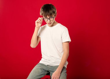 Stüdyoda parlak kırmızı arka planda poz veren beyaz tişörtlü gülümseyen genç çocuk, kendine güvenen, doğal duyguları olan sıradan bir gençlik portresi ve kopyalama alanı.