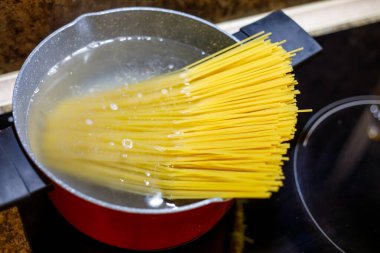 Makarna, mutfak sobasında kırmızı tencerede kaynayan suda pişiyor. İtalyan yemeği hazırlama süreci, ev yapımı yemekler, evde basit yemekler, konforlu yemekler ve günlük mutfak rutinleri..