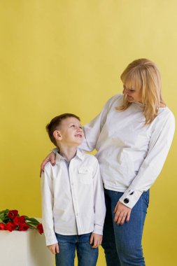 Anne ve küçük oğul sarı bir arka plana karşı birlikte duruyor, birbirlerine şefkatle bakıyor, sıcaklığı, güveni ve aile bağlarını ifade ediyorlar..