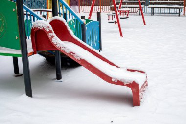 Boş kış parkında yoğun taze karla kaplı kırmızı oyun alanı kaydırağı. Soğuk kar yağışında parlak teçhizat beyaz zeminle çelişiyor
