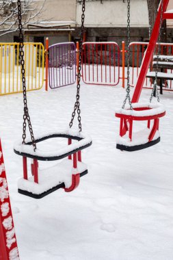 Kış parkında karla kaplı kırmızı çocuk salıncağına yakın dur. Soğuk mevsim havasında donmuş oyun alanı ekipmanları asılı duruyor