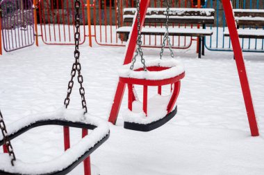 Kış parkında karla kaplı kırmızı çocuk salıncağına yakın dur. Soğuk mevsim havasında donmuş oyun alanı ekipmanları asılı duruyor