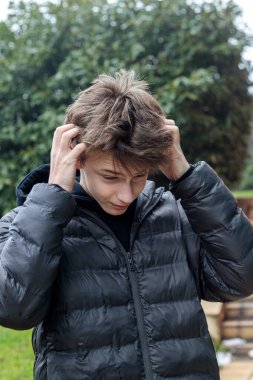 Genç çocuk dışarıda saçlarına dokunuyor, siyah kapüşonlu ve kabarık ceketli etkileyici ergen erkek kafası karışık ve düşünceli duygularını gösteriyor, parktaki modern gençliğin doğal samimi portresi.