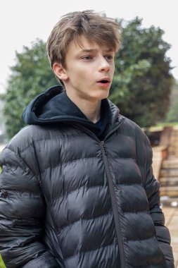 Genç çocuk şaşırmış bir ifadeyle dışarı bakıyor, siyah kapüşonlu ve kabarık ceketli genç bir erkek parkta duruyor, modern gençlik yaşam tarzının doğal bir portresi.