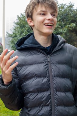 Gülümseyen genç çocuk parkta gülüyor. Siyah kapüşonlu ve kabarık ceketli, modern gençlik yaşam tarzının samimi duygusal portresi.