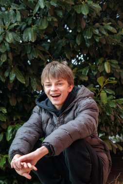 Siyah kapüşonlu ve kabarık ceketli gülümseyen bir gencin açık hava portresi, neşeli bir ifade ve otantik modern sokak gençlik yaşam tarzı.