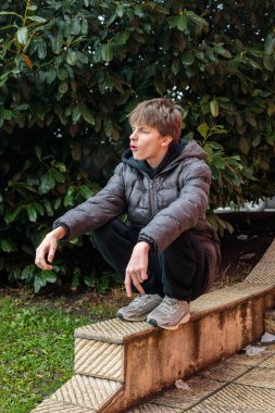 Parktaki kentsel basamaklara çömelmiş genç bir çocuğun açık hava portresi, anlamlı gençlik havası ve otantik modern sokak yaşam tarzı