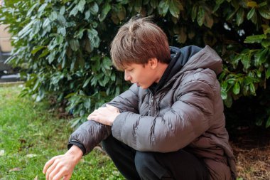 Açık havada kabarık ceketli bir gencin portresi parktaki ellerine bakıyor, düşünceli gençlik hali ve özgün modern yaşam tarzı konsepti.