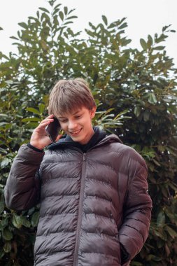 Açık hava portresi siyah kapüşonlu ve kabarık ceketli genç bir çocuk akıllı telefon, modern gençlik yaşam tarzı ve günlük dijital iletişim kullanıyor.