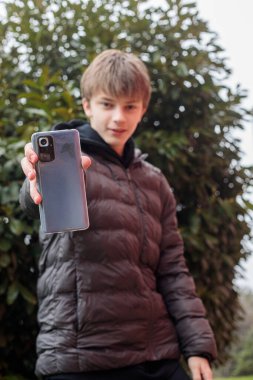 Şehir parkında akıllı telefonu kameraya, modern gençlik yaşam tarzına ve dijital iletişim konseptine doğru tutan bir gencin açık hava portresi