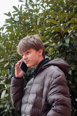 Açık hava portresi siyah kapüşonlu ve kabarık ceketli genç bir çocuk akıllı telefon, modern gençlik yaşam tarzı ve günlük dijital iletişim kullanıyor.