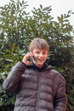 Açık hava portresi siyah kapüşonlu ve kabarık ceketli genç bir çocuk akıllı telefon, modern gençlik yaşam tarzı ve günlük dijital iletişim kullanıyor.