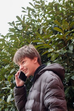 Açık hava portresi siyah kapüşonlu ve kabarık ceketli genç bir çocuk akıllı telefon, modern gençlik yaşam tarzı ve günlük dijital iletişim kullanıyor.