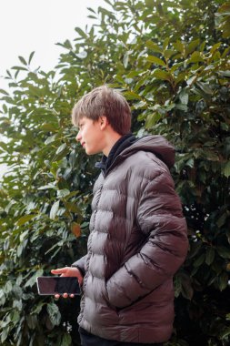 Açık hava portresi siyah kapüşonlu ve kabarık ceketli genç bir çocuk akıllı telefon, modern gençlik yaşam tarzı ve günlük dijital iletişim kullanıyor.