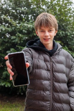 Ceketli bir gencin açık hava portresi elinde boş ekranlı akıllı telefon, modern gençlik yaşam tarzı ve dijital iletişim konsepti.