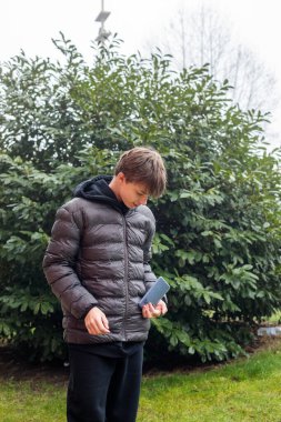 Açık hava portresi siyah kapüşonlu ve kabarık ceketli genç bir çocuk akıllı telefon, modern gençlik yaşam tarzı ve günlük dijital iletişim kullanıyor.