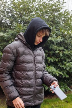 Açık hava portresi siyah kapüşonlu ve kabarık ceketli genç bir çocuk akıllı telefon, modern gençlik yaşam tarzı ve günlük dijital iletişim kullanıyor.