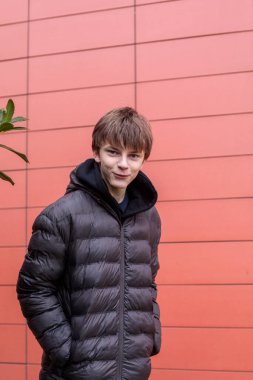 Siyah kabarık ceketli bir gencin modern kırmızı duvara bakarken kameraya baktığı portre. Kopya alanlı kentsel yaşam tarzı konsepti.