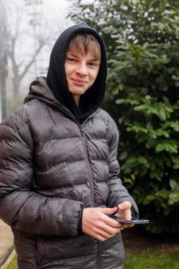 Siyah kapüşonlu ve kabarık ceketli gülümseyen bir gencin açık hava portresi, neşeli bir ifade ve otantik modern sokak gençlik yaşam tarzı.