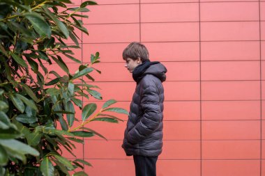 Kapüşonlu ceketli bir gencin modern kırmızı duvara karşı yeşil çalıların yanında duruşu, düşünceli ruh hali ve kopyalama alanı olan şehir manzarası.