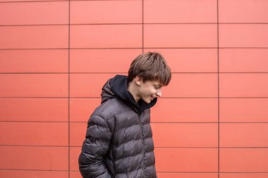 Siyah kabarık ceketli, gülümseyen genç bir çocuk modern kırmızı duvara bakarken yere bakıyor.