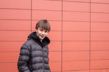 Siyah kabarık ceketli, kendine güvenen genç bir çocuk modern kırmızı duvara bakarken kameraya bakıyor.