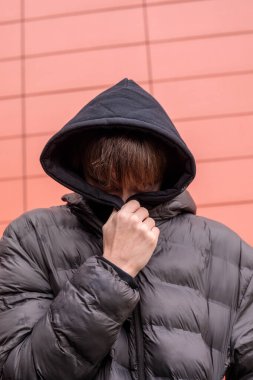 Siyah kapüşonlu ve kabarık ceketli bir gencin şehir portresi kırmızı duvarın yanında saklanan yüzü içe dönük ruh hali ve otantik modern sokak gençliği kavramı.