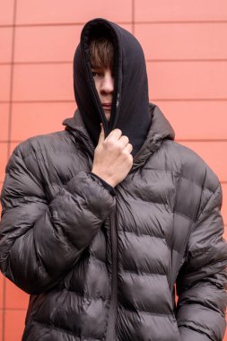 Siyah kapüşonlu ve kabarık ceketli bir gencin şehir portresi kırmızı duvarın yanında yüzünü kaplayan gizemli görünüş ve otantik sokak gençlik tarzı.