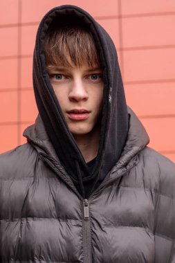 Kırmızı duvara karşı siyah kapüşonlu ve kabarık ceketli genç bir çocuğun portresi, yoğun bakışlar ve özgün modern sokak gençliği.