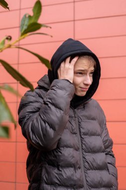 Kırmızı duvarın yanında siyah kapüşonlu ve kabarık ceketli genç bir çocuğun şehir portresi, duygusal ifade ve otantik modern sokak gençlik atmosferi.