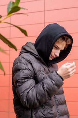 Kırmızı duvarın yanında siyah kapüşonlu ve kabarık ceketli genç bir çocuğun şehir portresi, odaklanmış ifade ve modern sokak tarzı gençlik hali.