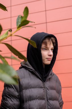Kırmızı duvarın yanında siyah kapüşonlu ve kabarık ceketli genç bir çocuğun şehir portresi, düşünceli bir ifade ve modern sokak tarzı gençlik atmosferi.