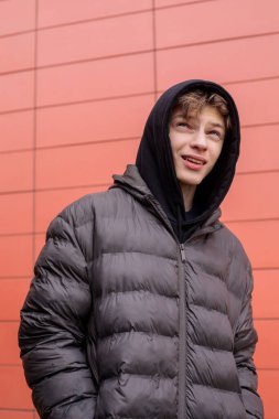 Kapüşonlu kabarık ceketli bir gencin portresini duvarlara yaslayın. Kafası karışmış bir gencin açık havada şüpheli bir ifadeyle yukarı baktığı samimi bir yaşam tarzı görüntüsü.