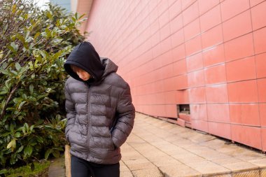 Kapüşonlu kabarık ceketli genç, fayanslı duvarlar boyunca yürüyor, modern kentsel alanda açık havaya bakan düşünceli ergen portresi.
